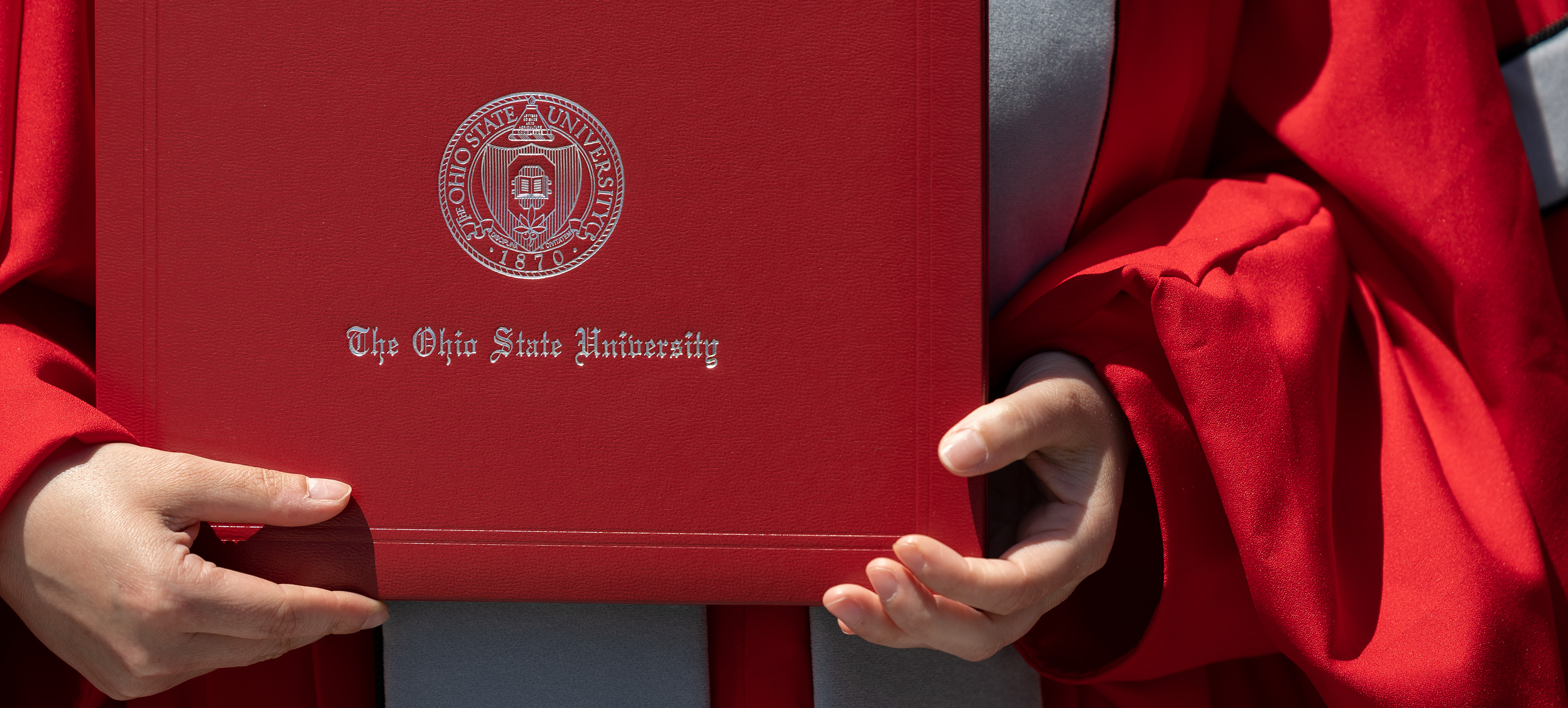 Phd student in scarlet and grey regalia holding a diploma binder