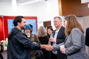 A gradaute student shacking hands with an Ohio State Dean introducing himsefl to her while another graduate student in back ground is drinking coffee and a second Dean joins as part of the 4 person conversation.