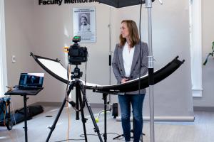 Graduate Student having a professional protrait taken during the appreciation week photo session. Student is stnading in front of backdrop while foreground shows camera equipment used to take the photo.