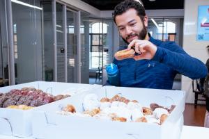 Graduate student picking up a glazed donut from the box of several varieties of donuts in the forground