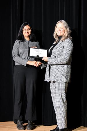 Mary Stomberger presenting the 1st place certificate to Ayushi Das on a stage