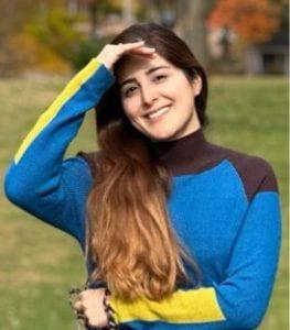 A photo of Elham Asghari Adib smiling wearing a blue, black, and yellow sweater with green foliage in the background.