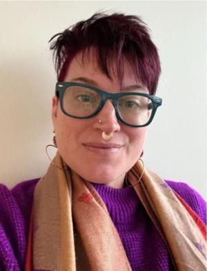 A photo of Audra Crouch smling at the camera wearing a purple sweater, scarf and glasses.