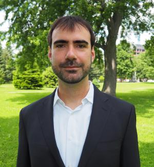 A photo of Enrico Berkes smiling wearing a white dress shirt, black blazer with green foliage in the background.