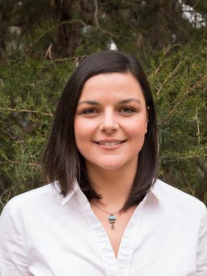 A photo of Jasmine Bruno smiling wearing a white dress shirt with green foliage in the background.