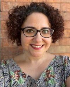 A photo of Burge Abiral smiling wearing a floral patterned blouse and glasses with a brick wall in the background.