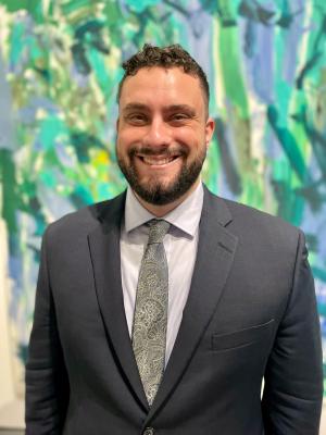 A photo of Adam Burston smiling wearing a grey suit and green tie with abstract art in the background.