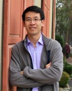 A photo of Marco Chen smiling with arms crossed wearing a purple dress shirt and grey sweater with an orange door in the background.