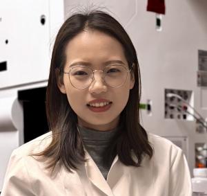 A photo of Caijao Dai smiling at the camera wearing glasses and a white blazer.
