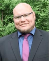 A photo of Andreas Fiedler smiling wearing glasses, a charcoal blazer, blue dress shirt, and pink tie with green foliage in the background.