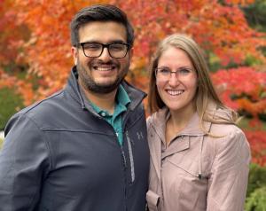 Varun and Tracy portrait in light jackets outside in front of a red leafed tree