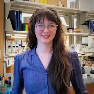 A photo of Rylee Hackley smiling at the camera wearing glasses and a blue dress with lab equipment in the background.
