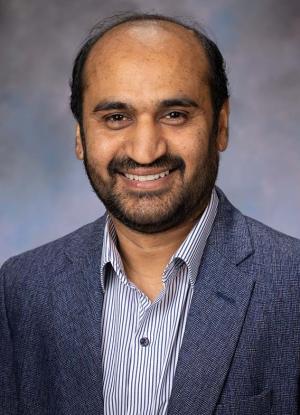A photo of Mubasher Hassan smiling wearing a grey-blue blazer and pinestripe dress shirt.