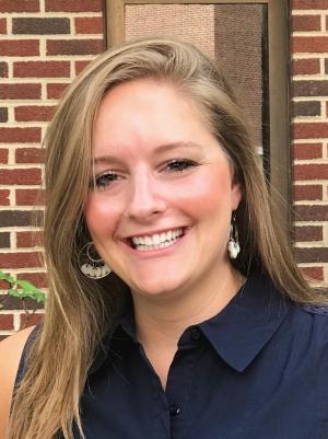 A photo of Laura Hildebrand smiling wearing a dark blue dres shirt with a brick wall in the background.