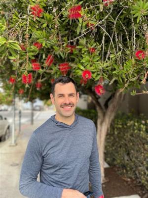 A picture of Jonathan Jacobs smiling at the camera in a blue shirt. Flowering tree in the background.