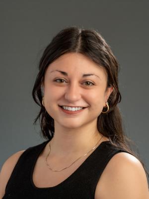 A photo of Kat Daiy smiling wearing a black dress with a grey background.