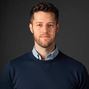 Photo of Daniel Nicholson smiling wearing a dark blue sweater over a blue dress shirt.