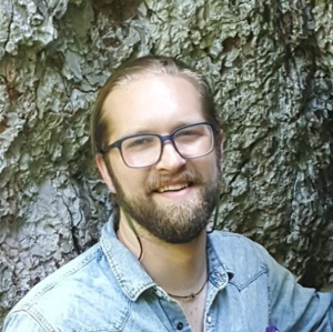 A photo of Shane Scaggs smiling at the camera wearing glasses and a denim button-up shirt with a natural rock formation in the background.