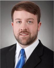 Joe Sharick smiling wearing a black blazer, white dress shirt, and blue tie.