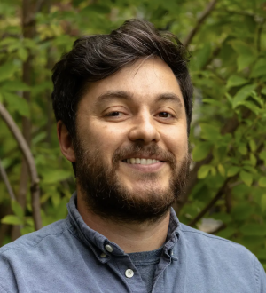 A photo of Sebastian Stockmaier smiling wearing a blue dress shirt with green foliage in the background.