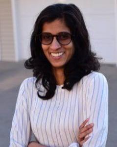 A photo of Swathy Krishna with her arms crossed smiling wearing a pinstripe white blouse and sunglasses.