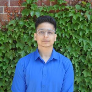 A photo of Mayank Tanwar wearing glasses and a blue dress shirt with a brick wall covered in green foliage in the background.