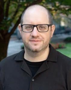 Photo of Adam Thomas smiling wearing glasses and a black dress shirt with green foliage in the background.