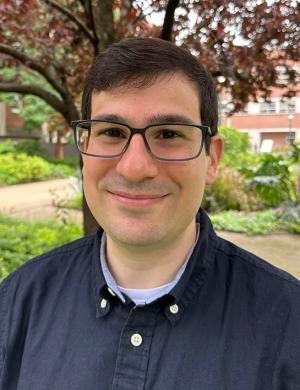 A photo of Marcus Vinicius Merfa smiling wearing a blue dress shirt with a tree and green foliage in the background.