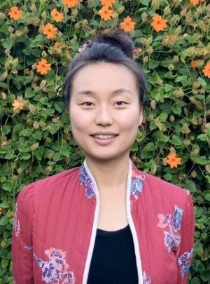 Grace Zhou smiling wearing a black blouse and red floral print blazer with green foliage and orange flowers in the background.