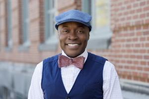 Stephen John Quaye wearing a blue hat and red bowtie in front of a brick wall